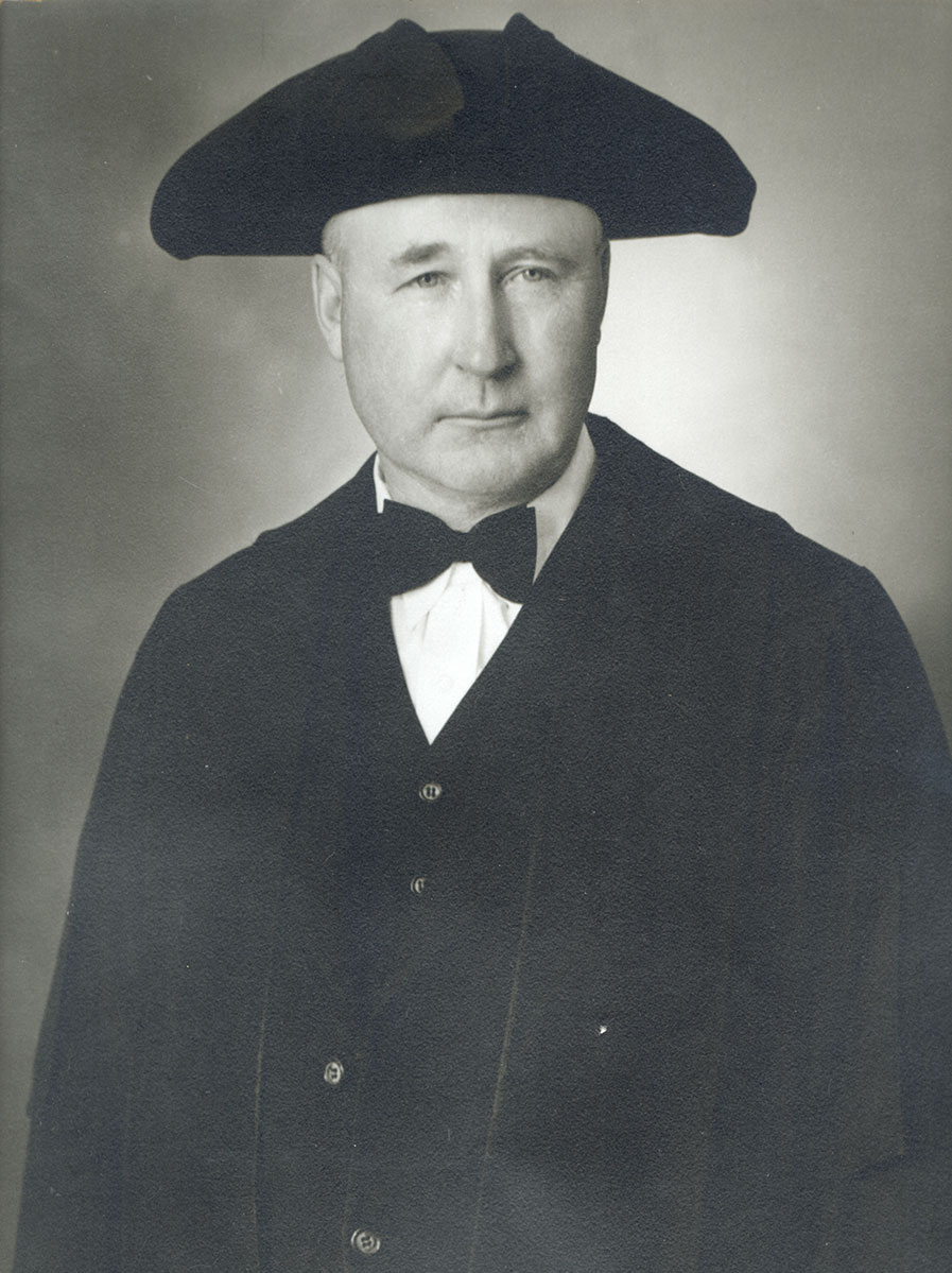 Black and white photo of white male in wardens cap and gown