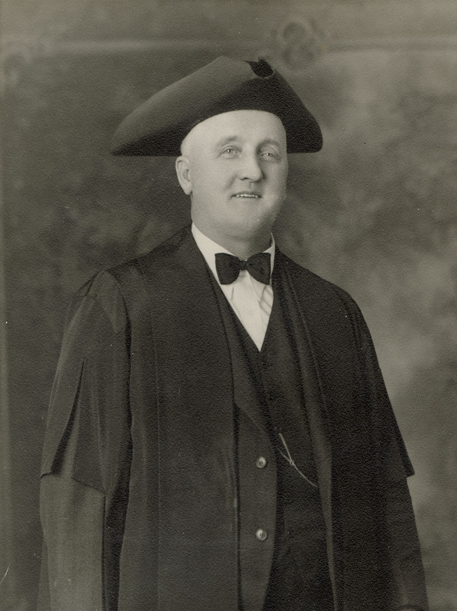 Black and white photo of white male in wardens cap and gown