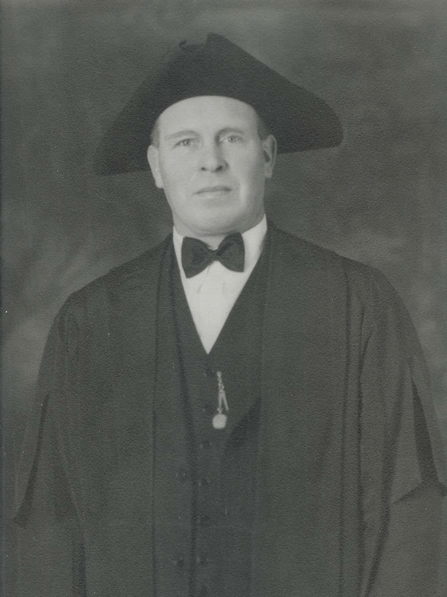 Black and white photo of white male in wardens cap and gown