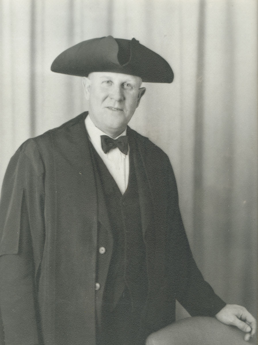 Black and white photo of white male in wardens cap and gown