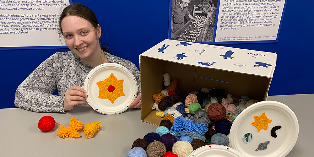 Lambton Heritage Museum staff Karissa Ramsey poses with an example yarn painting.