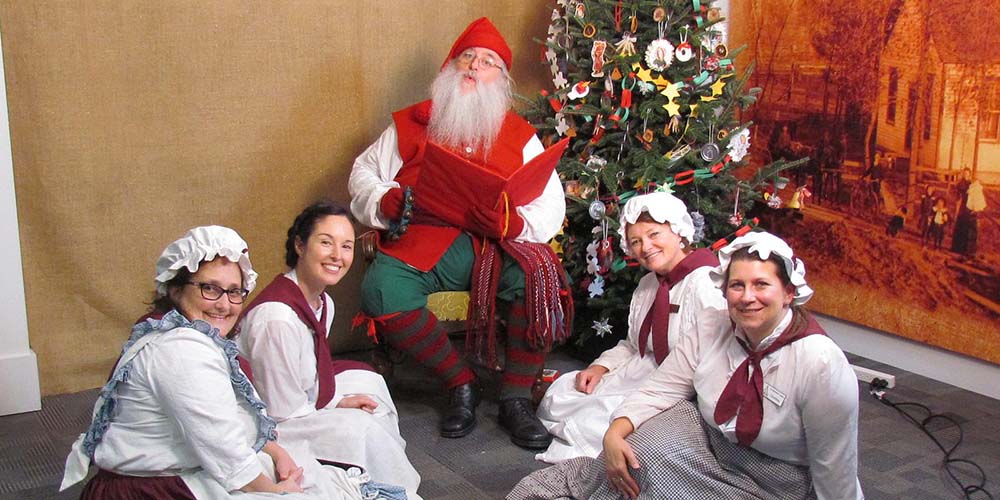 Santa Claus sitting in a chair with four Mrs. Claus around him.