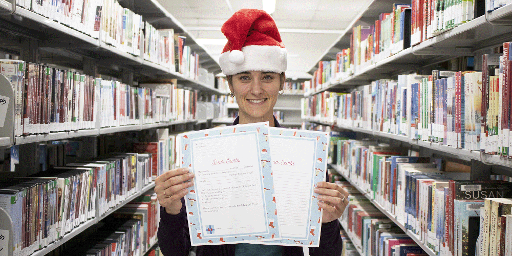 Lady standing between shelves of books holding out two papers, wearing a Santa hat.