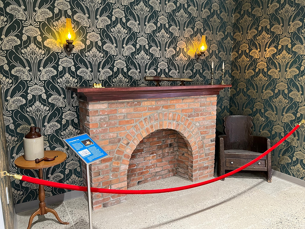 Brick fireplace against black and white wallpapered wall with light sconces behind velvet rope barrier.