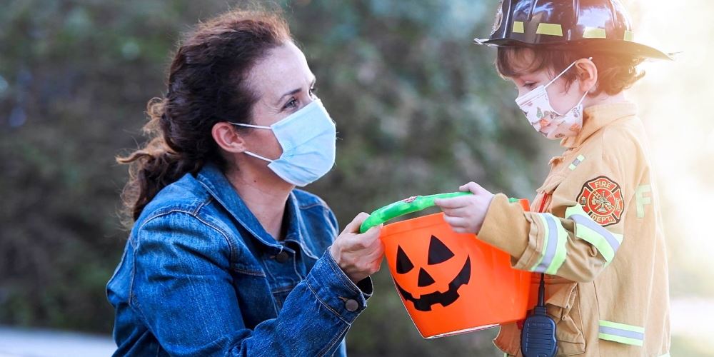 Woman wearing mask giving halloween pumpkin basket to child dressed as firefighter