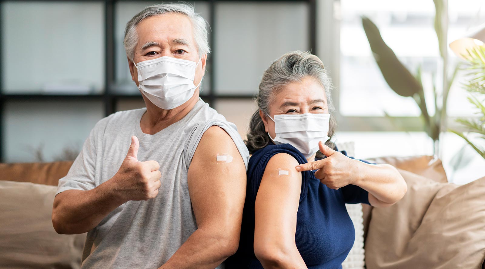 Senior man and woman wearing masks pointing at bandage on arms after vaccine
